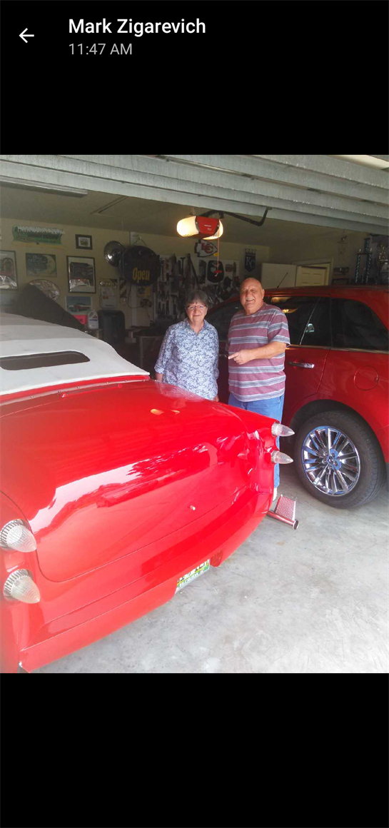 An older couple stands proudly next to a vintage red car in a cozy garage filled with memories.