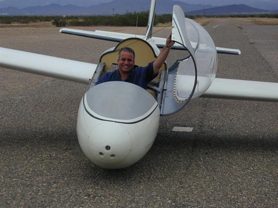 A pilot gives a thumbs-up while seated in a glider at an airstrip under blue skies.