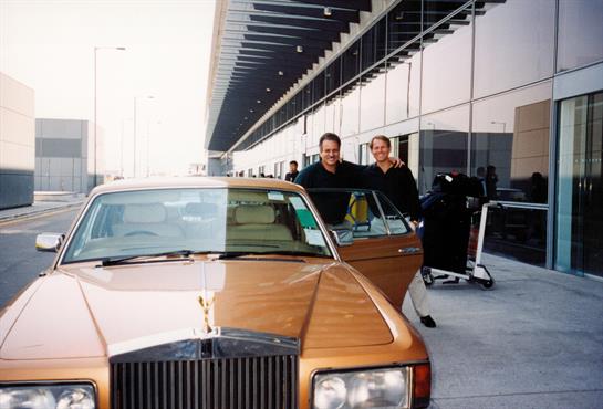 Two men stand beside a vintage car parked outside an airport terminal with modern architecture.