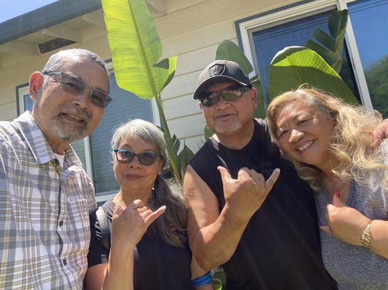 Four friends smile and pose with hand signs outside a house with tropical plants.