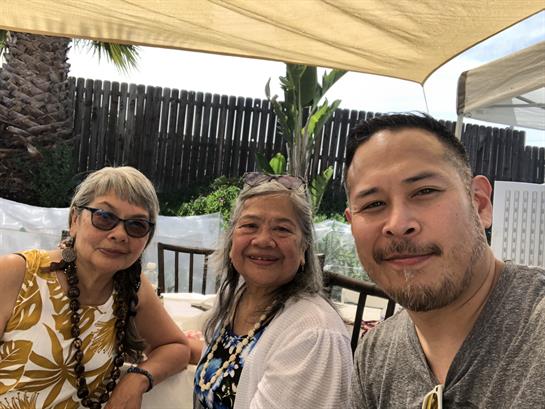 Three friends smile together under an outdoor canopy, enjoying a sunny day and each other’s company.