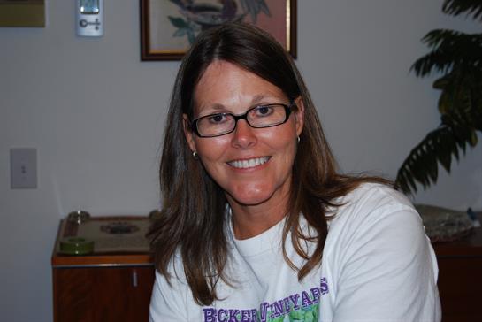 A cheerful woman with long hair and glasses smiles while sitting at a table, engaging with others.