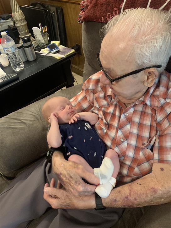 An elderly man enjoys quality time, holding a sleeping baby in a cozy chair at home.