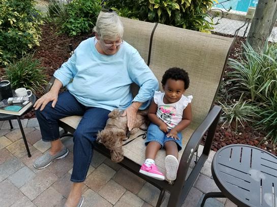 A senior woman and a young girl enjoy a sunny day sitting on a bench with a dog in a garden.
