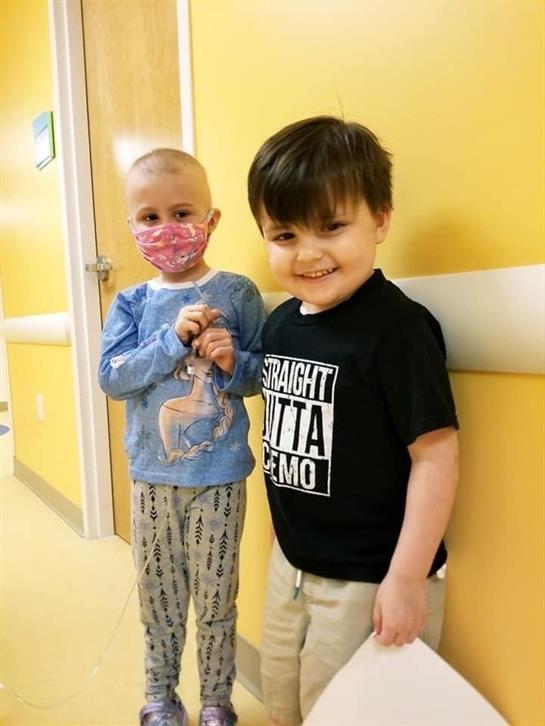 Two children stand in a bright hospital hallway, smiling and supporting each other during treatment.