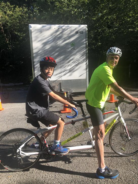 Two individuals enjoy a ride on a tandem bicycle, sporting helmets, in a park atmosphere.