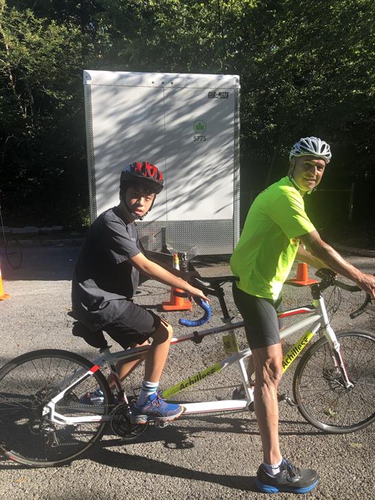 Two people ride a tandem bicycle along a trail with traffic cones in a park.