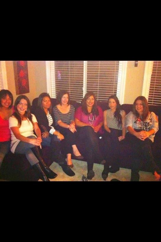 A group of seven friends sits together on a couch, chatting and smiling in a warmly lit living room.