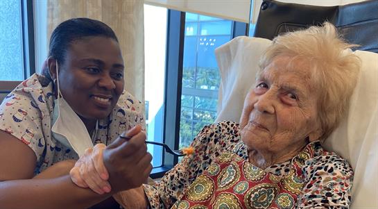 A caregiver smiles while feeding an elderly woman in a sunny room full of natural light.