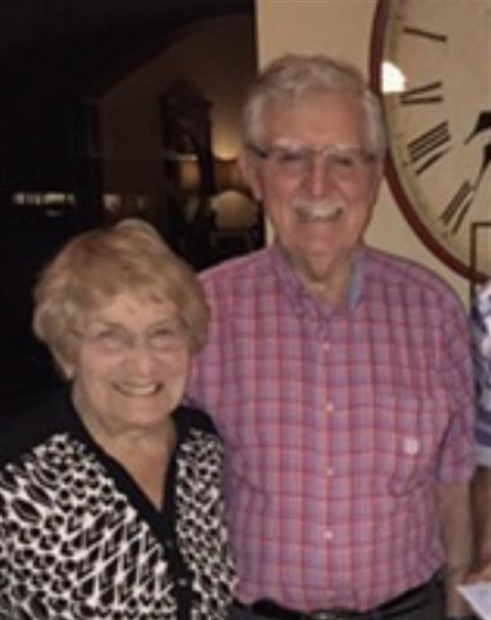 An elderly couple stands together, both smiling warmly in a cozy indoor setting with soft lighting.