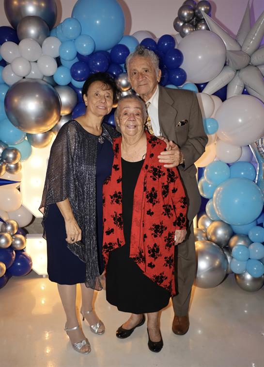 Three family members pose happily together against a backdrop of blue and silver balloons.