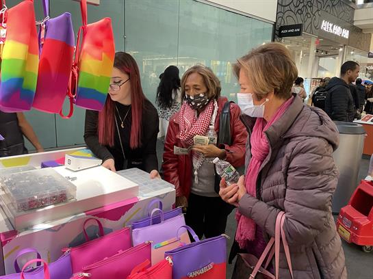 Three women actively browse a display of bright tote bags while enjoying their shopping day.