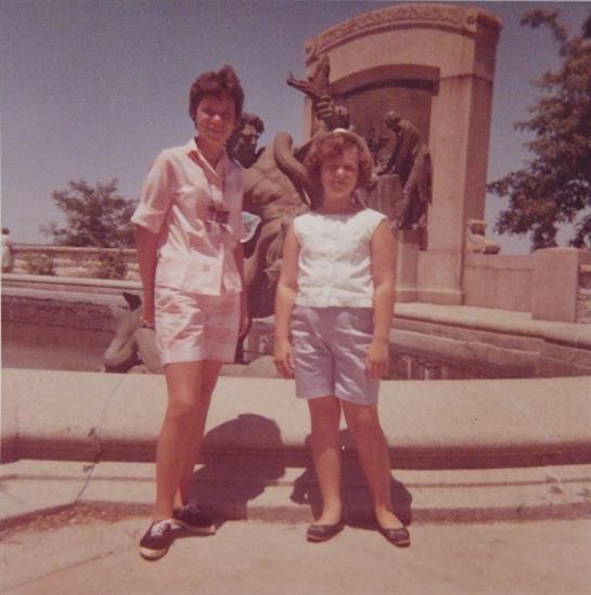 Two girls pose near a decorative fountain in a public park under clear skies.