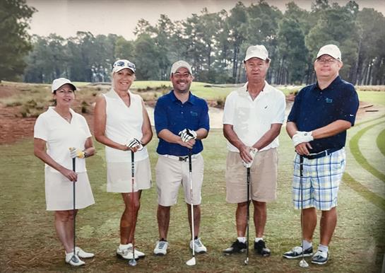 Six individuals stand on a golf course, smiling and holding clubs, enjoying their day outdoors.