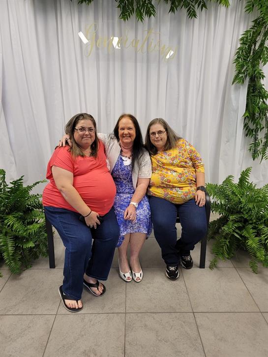 Three women sit closely together, smiling and enjoying a family moment surrounded by greenery.