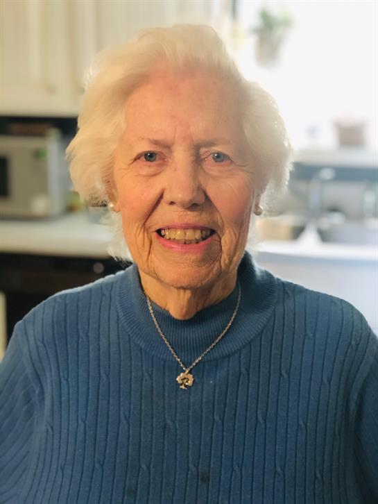 A smiling elderly woman in a blue sweater stands in a kitchen with white hair.