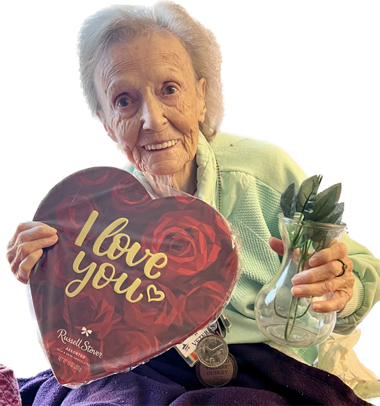 An elderly woman smiles while holding a heart-shaped box and a vase with flowers, celebrating love.