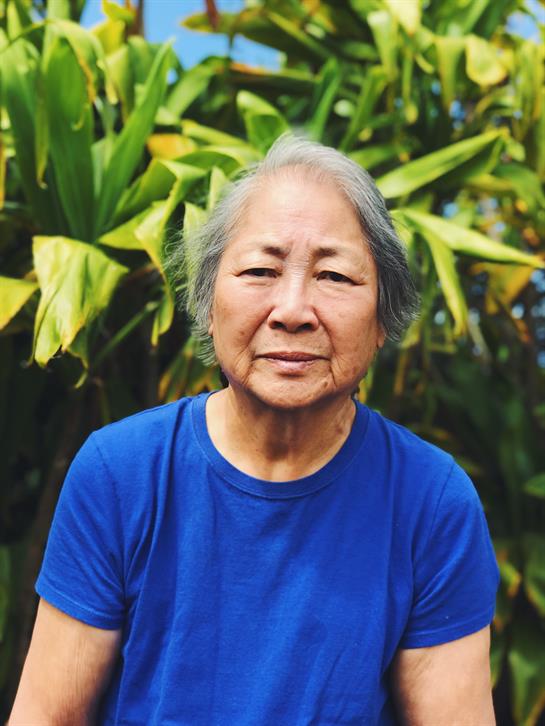 An elderly woman poses with a gentle smile surrounded by vibrant green plants in a garden.