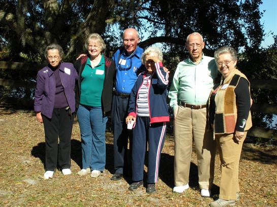 A group of six seniors stands happily outdoors, enjoying a social gathering in a park.