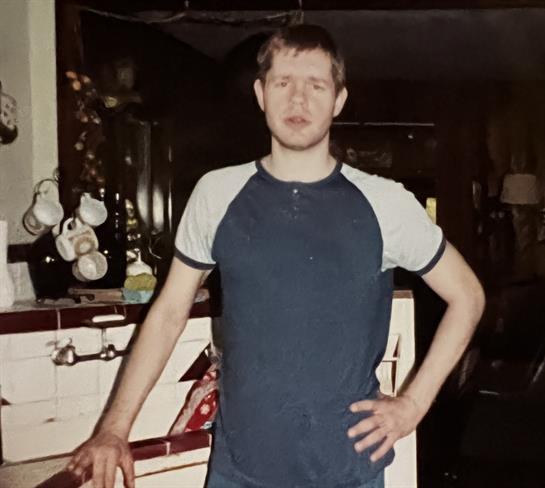 A young man poses with confidence in a vintage kitchen, wearing a blue shirt and jeans.