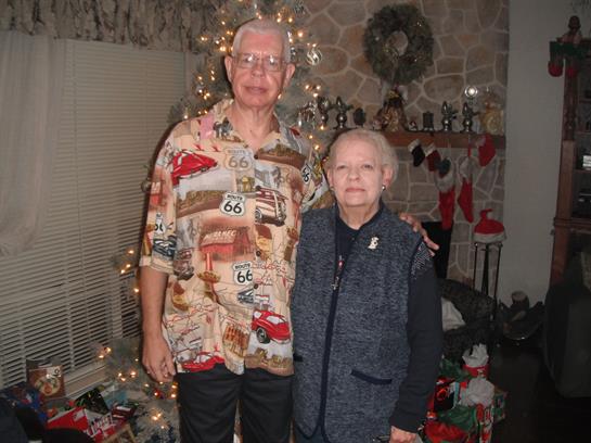 Couple stands smiling in a warmly decorated living room with holiday decorations and lights.