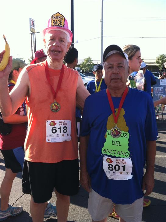 Two older men pose proudly after completing a running event, showing off their medals and smiles.