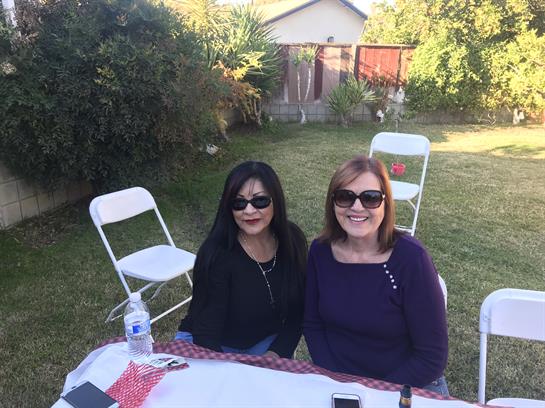 Two women sit at a table outdoors, smiling and enjoying their time in a bright garden setting.