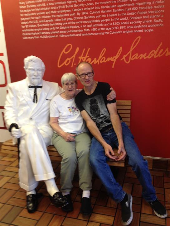 Two visitors sit together with a white-suited statue in a casual restaurant atmosphere.