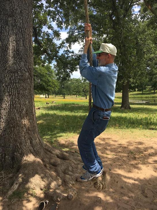 A man wearing casual clothes climbs a rope attached to a tree in a park surrounded by greenery.