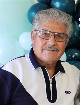 Elderly man with glasses smiles warmly at a lively gathering decorated with balloons.
