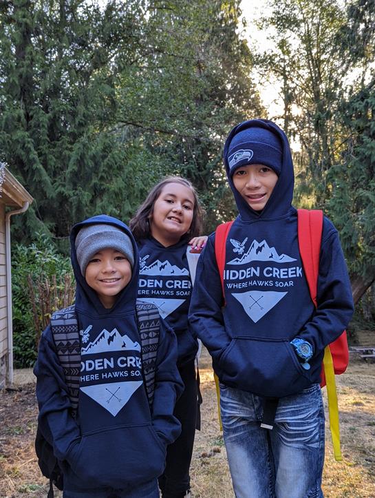 Three children pose happily in matching hoodies, surrounded by trees on a sunny afternoon.