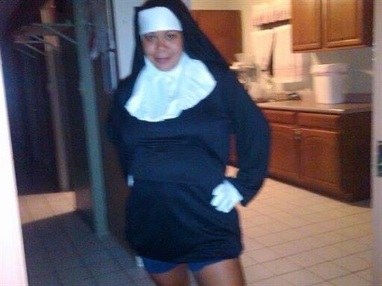 Woman dressed in humorous nun outfit stands proudly in a cozy kitchen during a fun party.