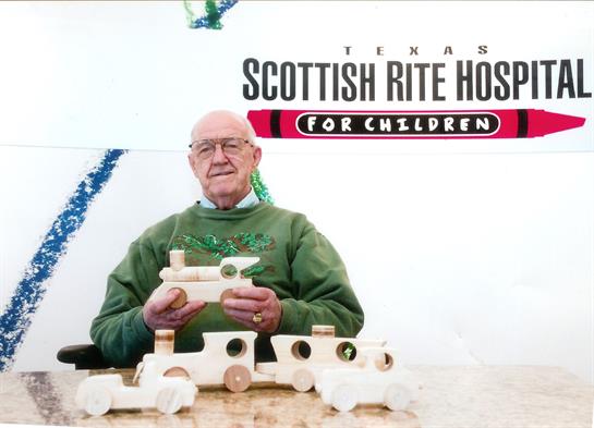 An elderly man proudly displays several handmade wooden toy cars at a children's hospital event.