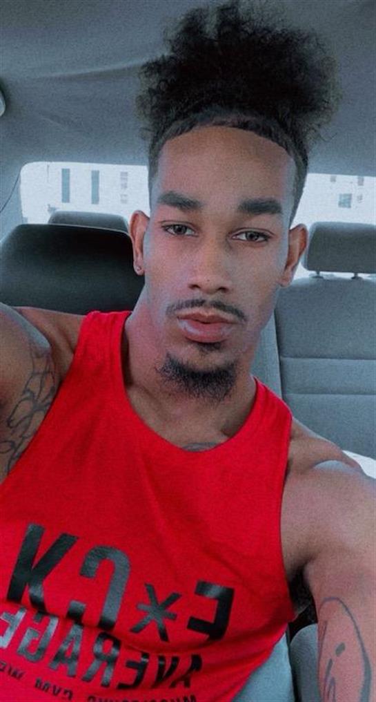 A young man takes a selfie in a car, wearing a red tank top and showcasing his tattoos.