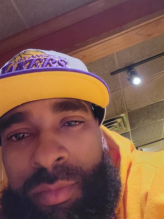 Close-up of a man with a beard wearing a bright yellow Lakers cap and a matching hoodie, smiling.