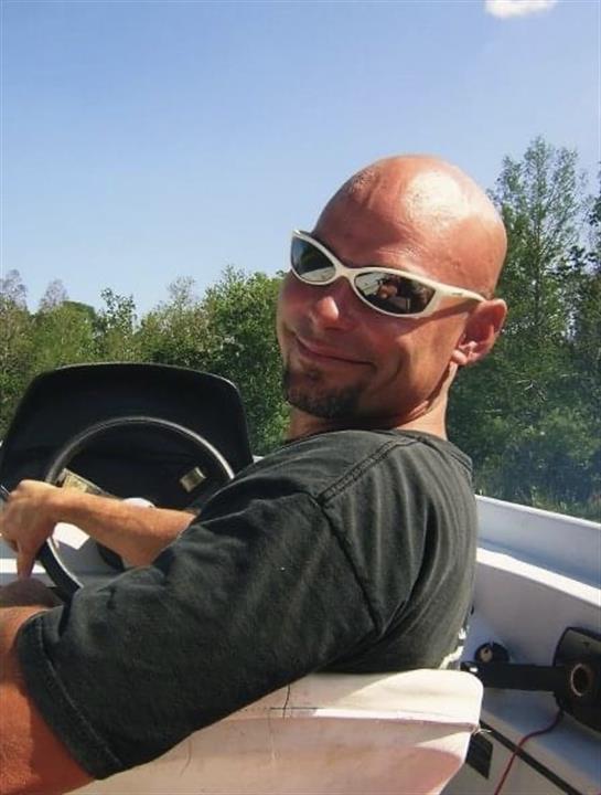 Man enjoys a sunny day driving a boat through a scenic waterway with trees nearby.