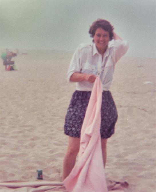 A woman smiles while holding a blanket on a sandy beach, ready to relax under the sun.