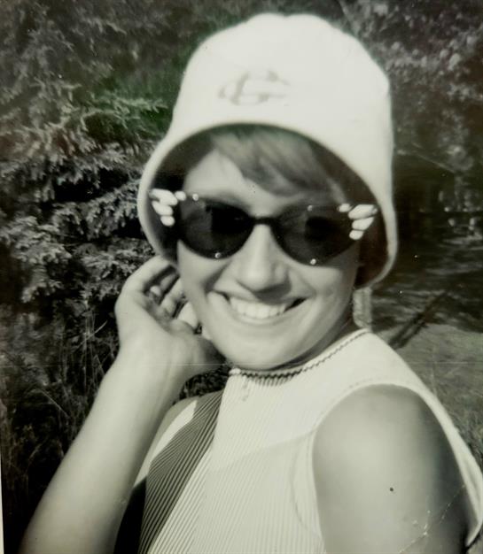 A woman smiles brightly while posing outdoors, showcasing her fashionable sunglasses and hat.