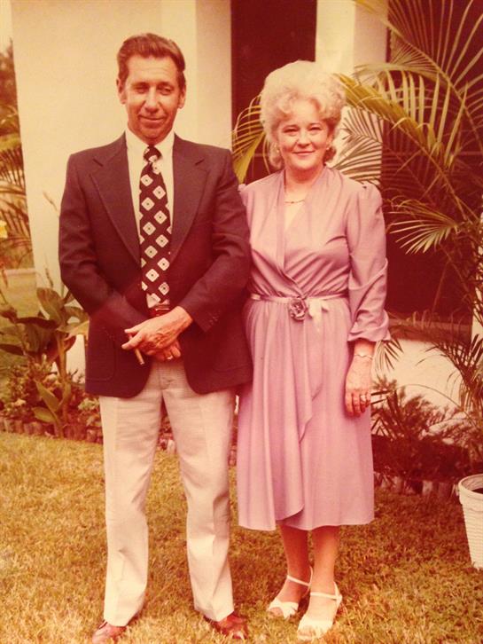 A man and woman stand side by side, smiling in a lush garden with tropical plants.