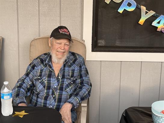 A senior man with a long beard sits cheerfully in a chair, adorned with festive decorations.