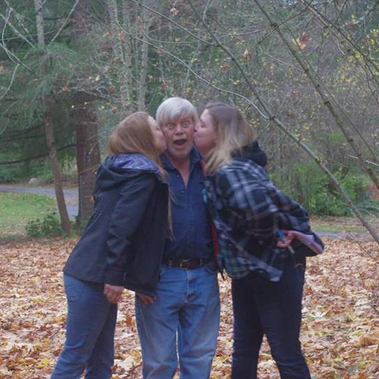 Two women kiss a man playfully in a leaf-covered forest, enjoying a lighthearted gathering.