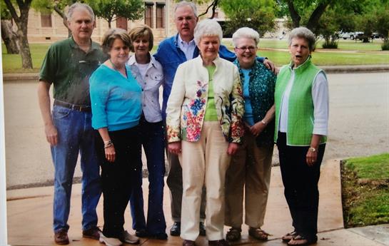 A cheerful gathering of friends in bright clothing smiles and poses for a group photo outdoors.