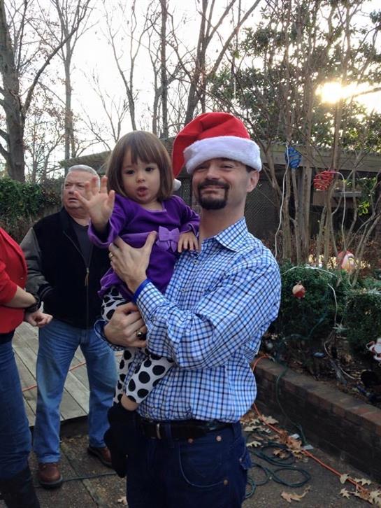 A joyful holiday gathering shows a man and a girl in a Santa hat, with others behind them.
