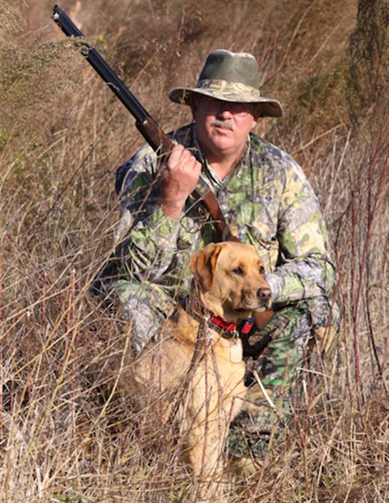 A man in camouflage holds a rifle while kneeling next to a golden retriever in tall grass.