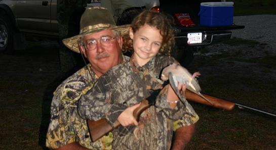 A man and a young girl smile proudly while holding a small bird and a hunting rifle outdoors.