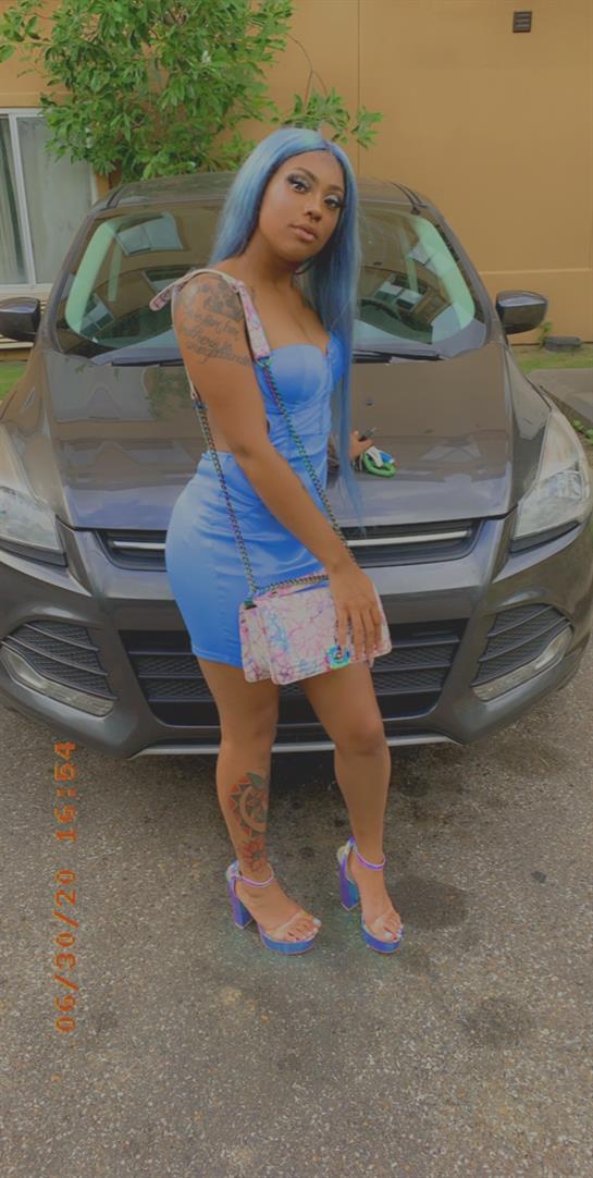 A woman stands confidently in a blue dress, showcasing her style near a gray SUV under sunlight.