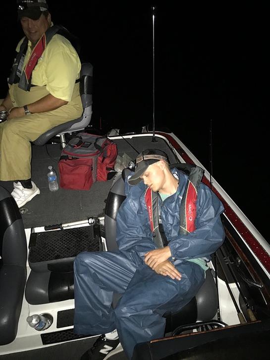 Two people are on a boat in the dark, one resting while the other operates the controls.