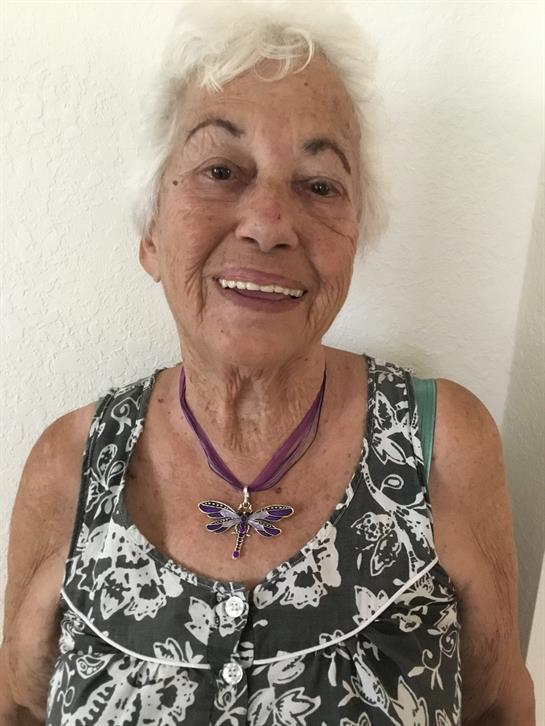 Elderly woman smiles by a light wall, showcasing her unique necklace and dress.