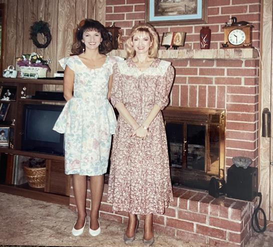 Two women stand side by side, smiling, in a decorated living room with a brick fireplace.