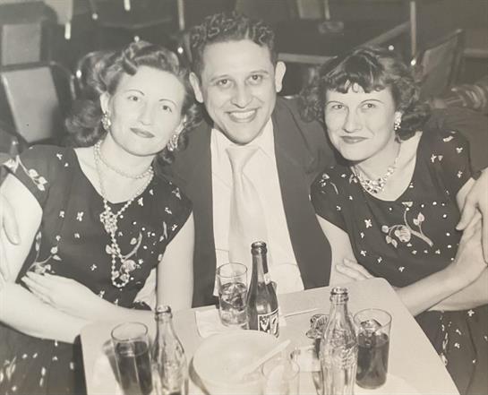 Three friends share smiles and drinks at a gathering, dressed in stylish outfits from the 1950s.